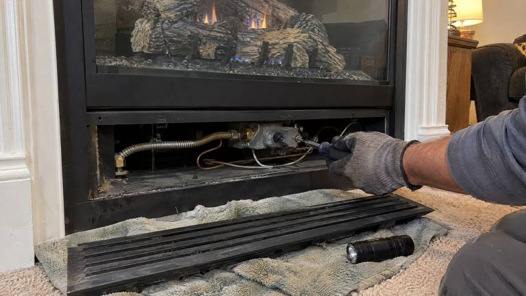 A gloved hand works on the gas valve behind the removed lower louver of a gas fireplace, with the panel and a flashlight on a towel.