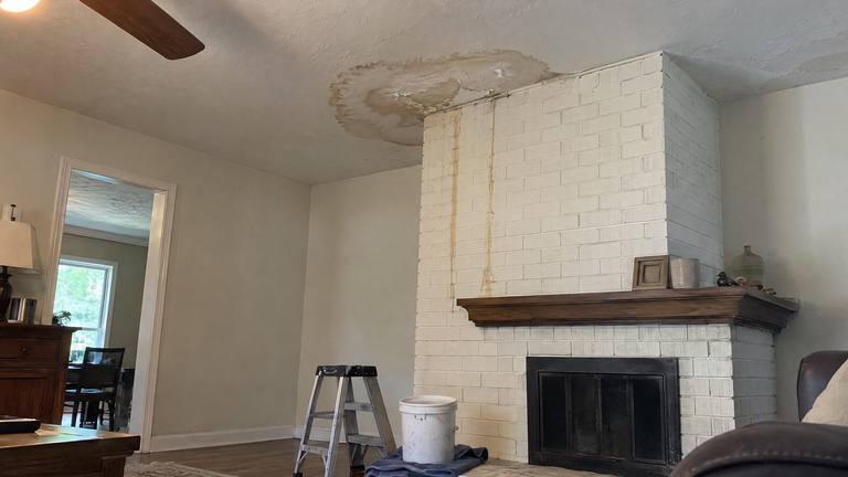 A brown water stain spreads across a ceiling beside a painted brick chimney breast, with drip marks on the brick and a bucket below.